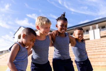 Smiling multiracial elementary boys standing with arm around against school building on sunny day