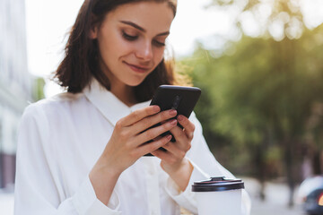 Close up of hands using cell phone with background of the city, smiling girl texting with friends while walking outdoor