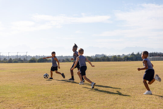 Multiracial elementary schoolboys playing soccer on school soccer field against sky during sunny day