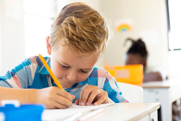 Close-up of cute caucasian elementary schoolboy writing on book while sitting at desk in school