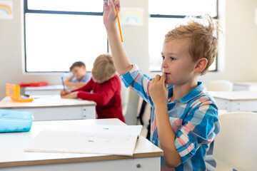 Caucasian elementary schoolboy raising hand while sitting at desk in classroom