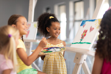 Cheerful multiracial elementary schoolgirls standing by caucasian girl painting on easel in class
