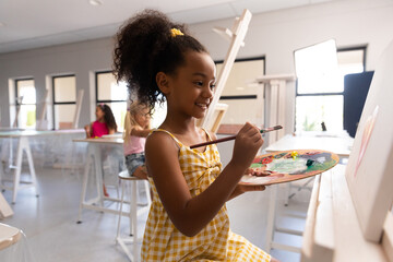 Smiling biracial elementary schoolgirl with palette and paintbrush painting on easel in school