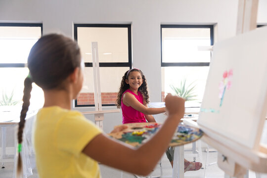 Smiling multiracial elementary schoolgirls painting on easel during drawing class in school