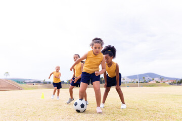 Full length of multiracial elementary schoolgirls in sports uniform playing soccer against sky