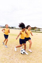 Full length of multiracial elementary schoolgirls playing soccer on school ground against sky