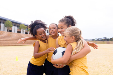 Fototapeta Cheerful multiracial elementary schoolgirls with soccer ball embracing while standing on ground obraz