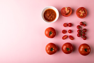 Overhead view of fresh puree in bowl by various red tomatoes by copy space on pink background