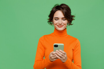 Young smiling happy woman 20s wear casual orange turtleneck hold in hand use mobile cell phone browsing internet isolated on plain pastel light green color background studio People lifestyle concept. - obrazy, fototapety, plakaty