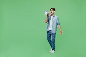 Full size body length side view promoter young brunet man 20s years old wears blue shirt hold scream in megaphone announces discounts sale Hurry up isolated on plain green background studio portrait.