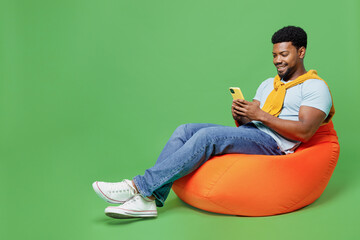 Full body young man of African American ethnicity 20s in blue t-shirt sit in bag chair hold in hand...