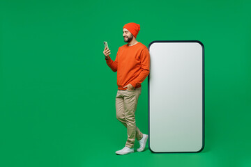 Full body young smiling man 20s wear orange sweatshirt hat stand near big blank screen mobile cell phone with workspace copy space mockup area use smartphone isolated on plain green background studio.