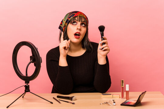 Young makeup artist woman doing a makeup tutorial isolated on pink background pointing upside with opened mouth.