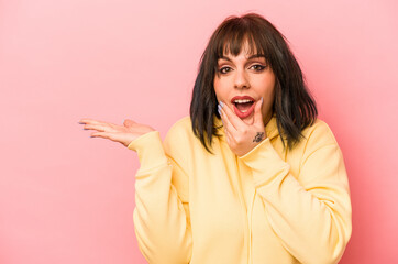 Young caucasian woman isolated on pink background holds copy space on a palm, keep hand over cheek. Amazed and delighted.