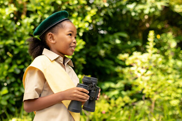 Smiling african american scout girl in uniform holding binoculars in forest - Powered by Adobe