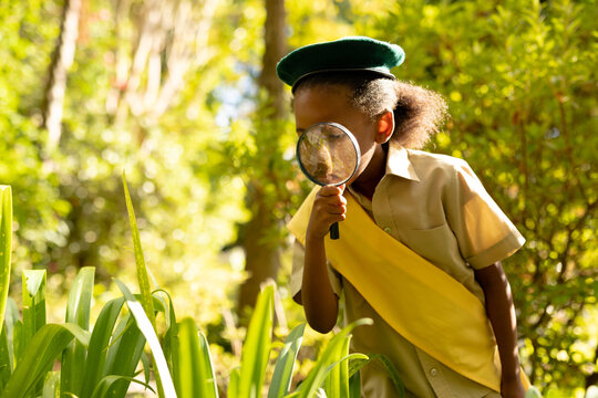 African american scout girl in uniform looking at plants through magnifying glass in forest