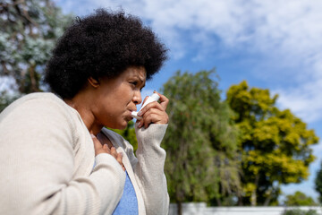 African american mid adult woman with hand on chest using asthma inhaler while standing outdoors - obrazy, fototapety, plakaty