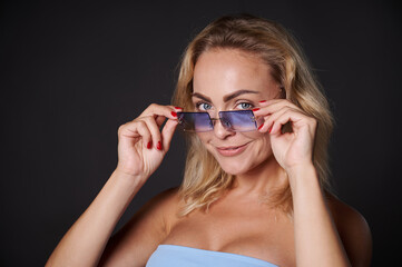 Naklejka premium Headshot beautiful blonde European woman confidently looking at camera through blue sunglasses, isolated over black background with copy space for ads