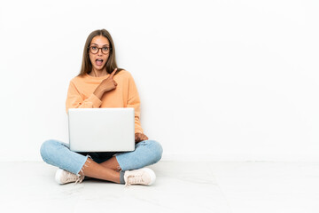 Young woman with a laptop sitting on the floor surprised and pointing side