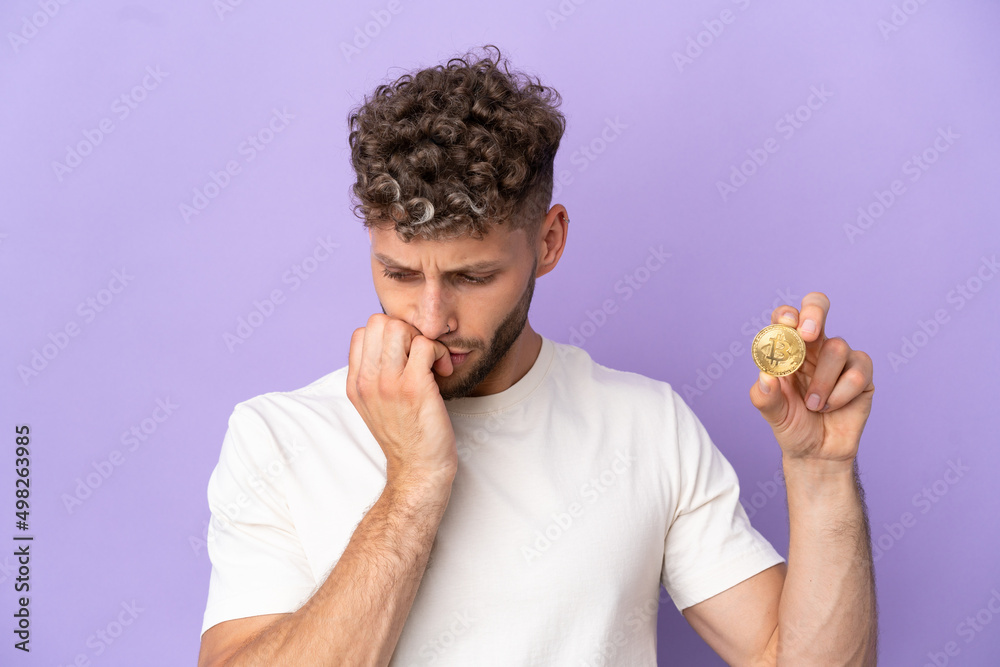 Wall mural Young caucasian man holding a Bitcoin isolated on purple background having doubts