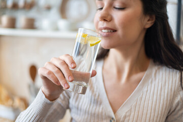 Refreshment concept. Young woman drinking water with lemon, holding glass, standing in kitchen, closeup