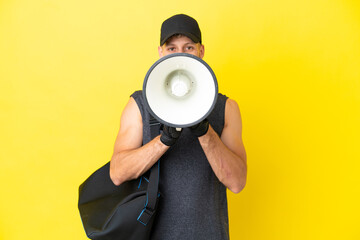 Young sport blonde man with sport bag isolated on yellow background shouting through a megaphone