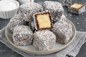 Traditional Australian Lamington cake in chocolate glaze and coconut flakes on a plate on a gray concrete background with a cup of tea. Selective focus.