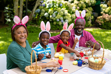 Fototapeta premium Portrait of happy african american siblings and grandparents in bunny ears painting easter eggs