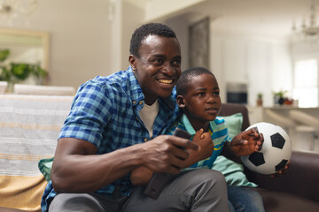 Happy african american boy and father with soccer ball sitting on sofa at home