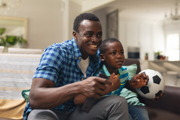 Happy african american boy and father with soccer ball sitting on sofa at home - Powered by Adobe