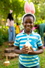 Fototapeta premium Portrait of smiling african american boy in bunny ears holding easter egg while family in backyard