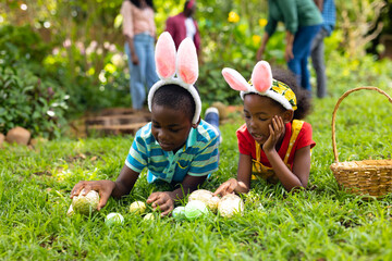 Naklejka premium African american siblings in bunny ears arranging easter eggs on grass while family in background