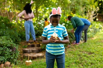 Fototapeta premium Happy african american boy in bunny ears looking at easter egg while family searching in backyard