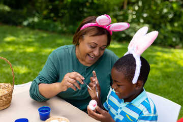 Smiling african american boy and grandmother in bunny ears painting egg at backyard on easter day