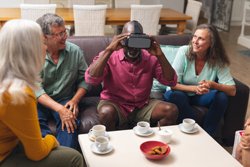 Happy multiracial senior male and females looking at friend using vr headset at home