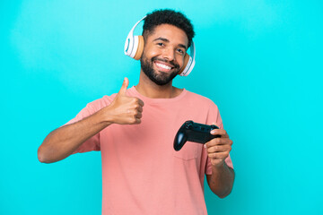 Naklejka premium Young brazilian man playing with a video game controller isolated on blue background giving a thumbs up gesture