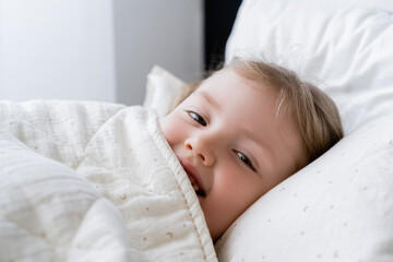 Fototapeta na wymiar cheerful child looking at camera while lying in bed under white blanket.