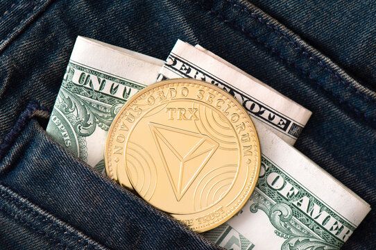 A golden physical TRON coin and folded dollars sticks out of the pocket of black jeans close-up. Saving and exchange money in cryptocurrency concept. Business background.