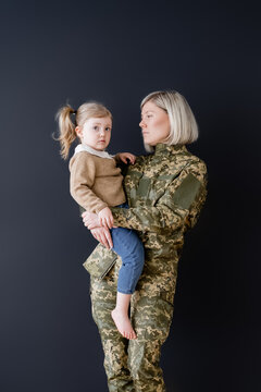 serious woman in camouflage holding little daughter in hands isolated on black.