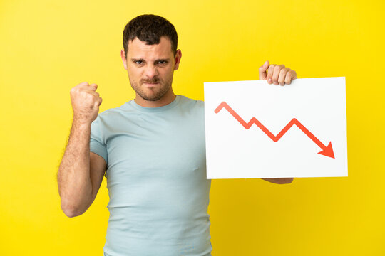 Brazilian man over isolated purple background holding a sign with a decreasing statistics arrow symbol and angry