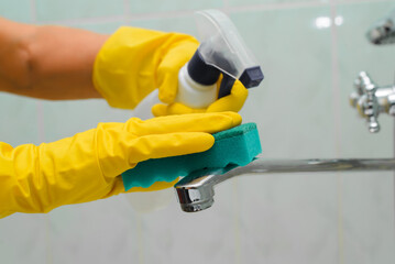 Hygiene, bathroom cleaning. Close-up of hands in rubber gloves cleaning water faucet, disinfecting with chemical spray, detergent. Selective focus on hand with sponge