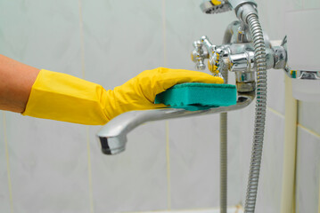 Cleaning bathroom, hand in yellow rubber glove washing chrome faucet with sponge. Close-up, selective focus