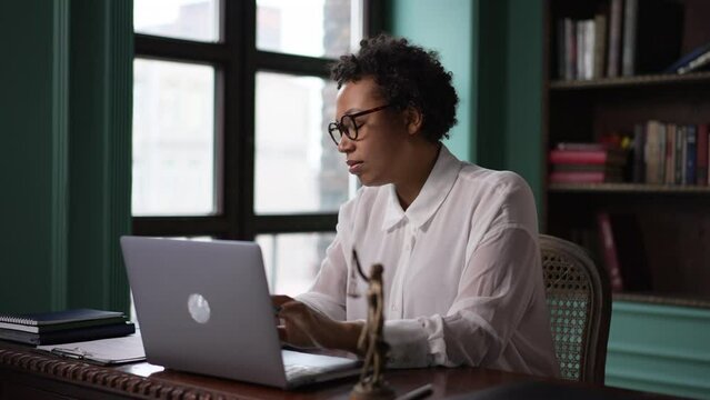 Young businesswoman or lawyer working with laptop and sitting at workplace with law symbol spbas. 4k Portrait of american african woman looks at suit and types on device keyboard, does routine work