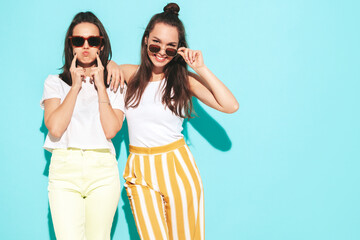 Two young beautiful smiling blond hipster female in trendy summer clothes. Sexy carefree women posing near blue wall in studio. Positive models having fun. Cheerful and happy. In sunglasses