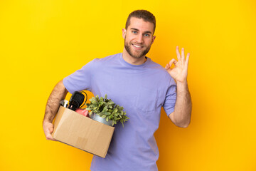 Young caucasian making a move while picking up a box full of things isolated on yellow background...