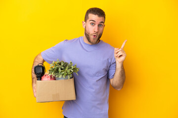 Young caucasian making a move while picking up a box full of things isolated on yellow background...