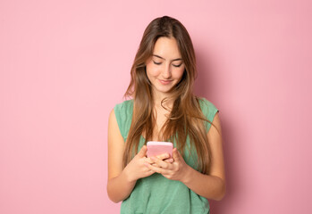Portrait of a smiling young casual caucasian woman looking at mobile phone isolated over pink background
