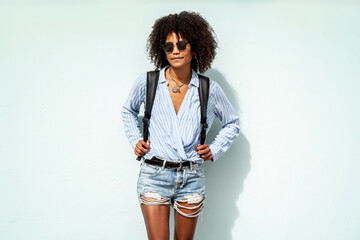 Fashionable woman with afro hairstyle posing on the city wall, wearing short jeans, shirt,...