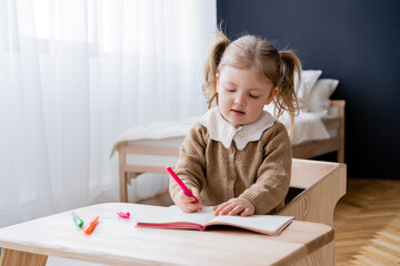Naklejka premium little girl drawing with colorful felt pens while sitting at table in bedroom.