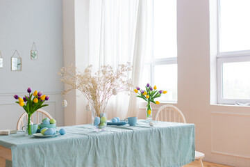 Spacious bright room with a table decorated with Easter eggs and bouquets of flowers and a blue tablecloth in daylight.